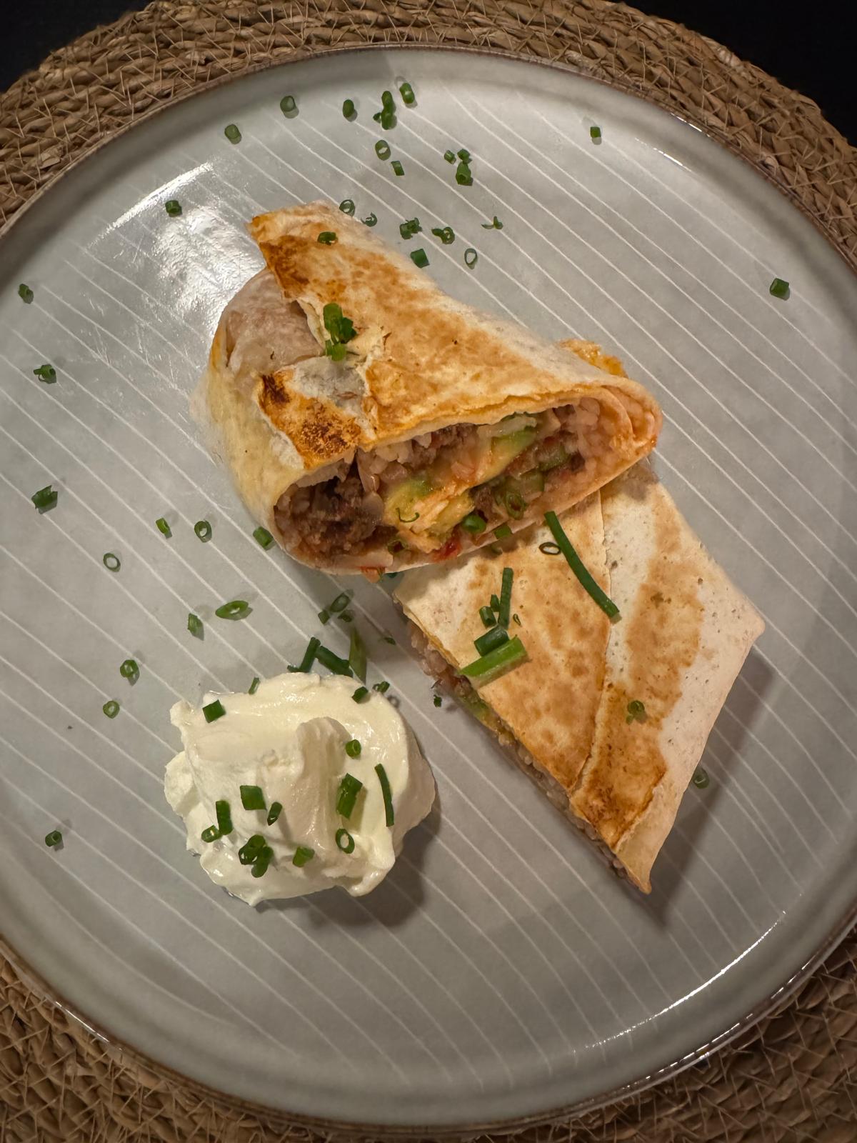 Aufgeschnittener Burrito mit Tomatenhackfüllung auf einem grauen Teller, mit Schmand und Frühlingszwiebeln